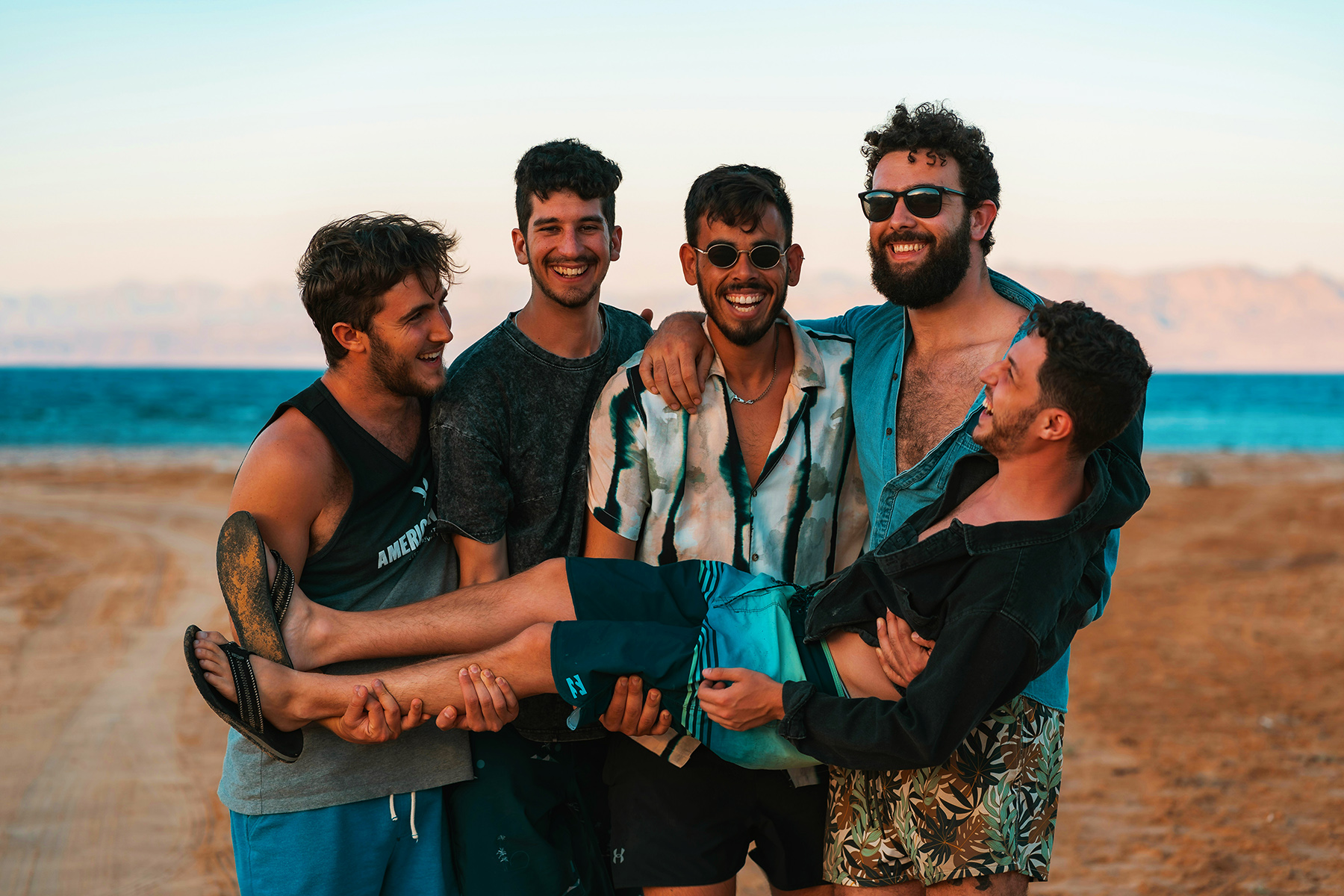 A group of queer men laughing together by the sea at sunset.